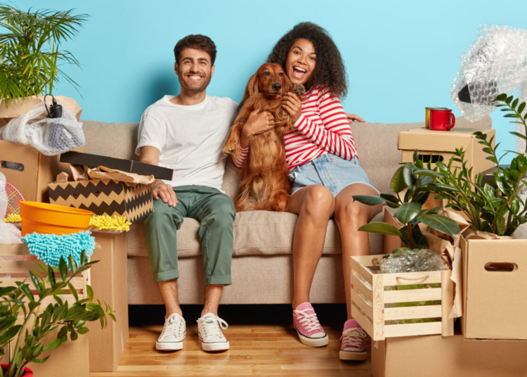 Relocation, renovation and removal concept. Happy diverse couple sit on sofa with favourite dog, have break on moving day, busy unpacking, have mess in new flat, many packages, remove to own house