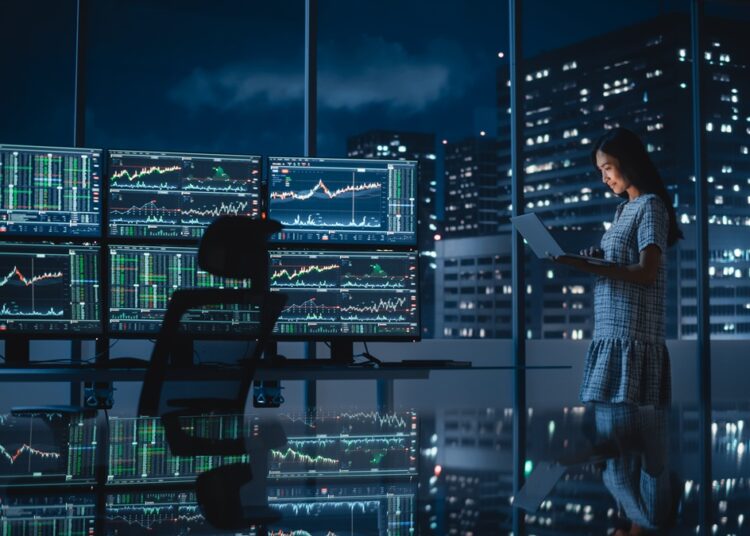 Financial Analyst Using Laptop Computer, Standing Next to Multi-Monitor Workstation with Real-Time Stocks, Commodities and Exchange Market Charts. Businesswoman Working in Investment Bank City Office.