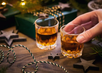 Woman hand holds a Glass of whiskey or bourbon with Christmas decoration on dark background. New Year, Christmas and winter holidays whiskey mood concept