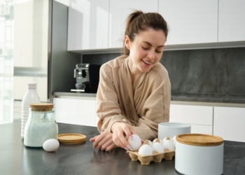 Attractive young cheerful girl baking at the kitchen, making dough, holding recipe book, having ideas.