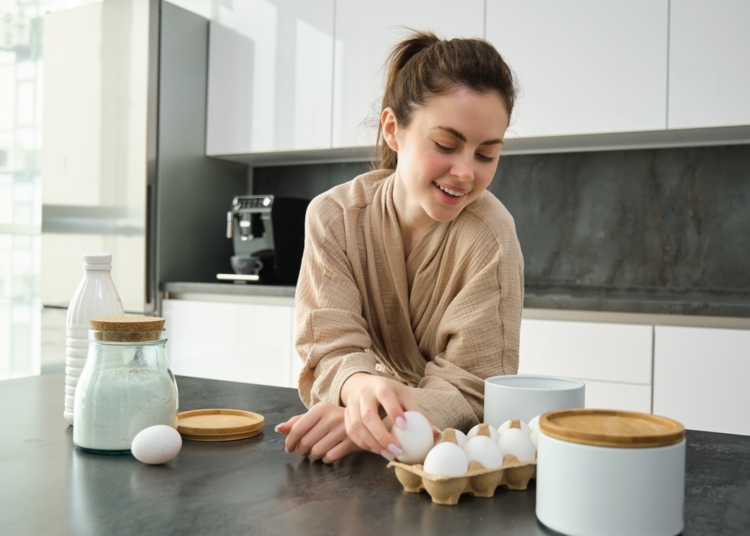Attractive young cheerful girl baking at the kitchen, making dough, holding recipe book, having ideas.